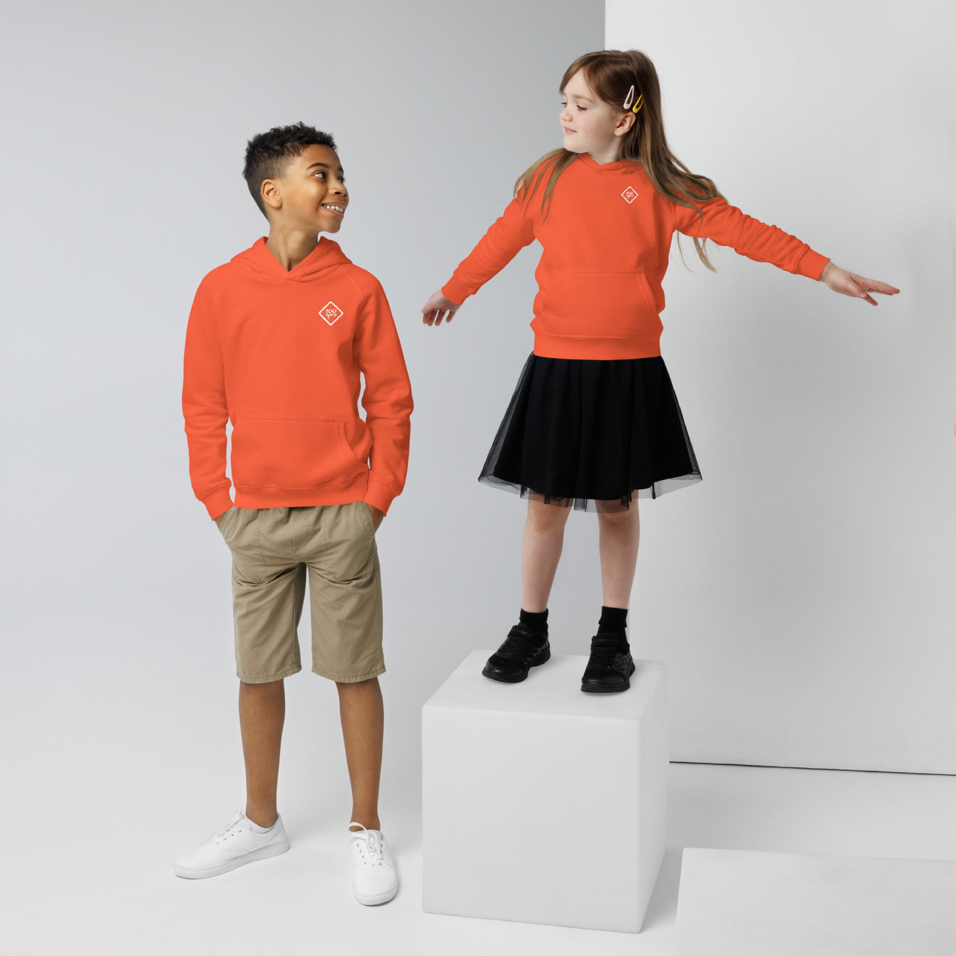 Two kids wearing the "Unisex Kids Eco Hoodie | 100yrs Logo" in vibrant orange; one wearing shorts and the other a skirt.