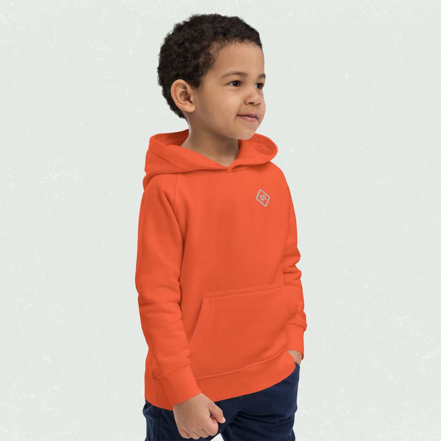 Young child wearing an orange Unisex Kids Eco Hoodie with the 100yrs logo, featuring a front pouch pocket and a cozy hood.