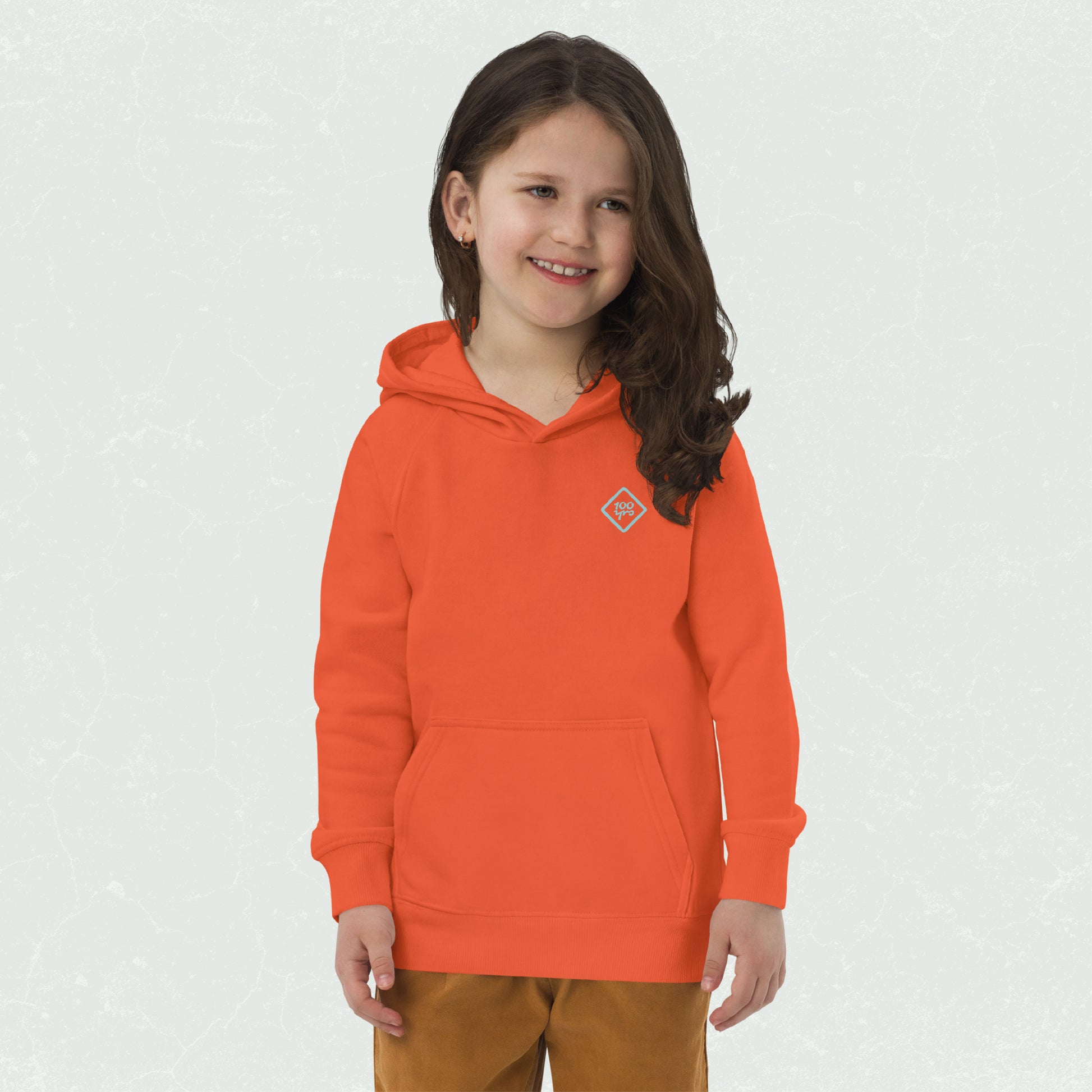 A smiling child wearing a bright orange unisex eco-friendly hoodie with a 100yrs logo, featuring a front pouch pocket and raglan sleeves.