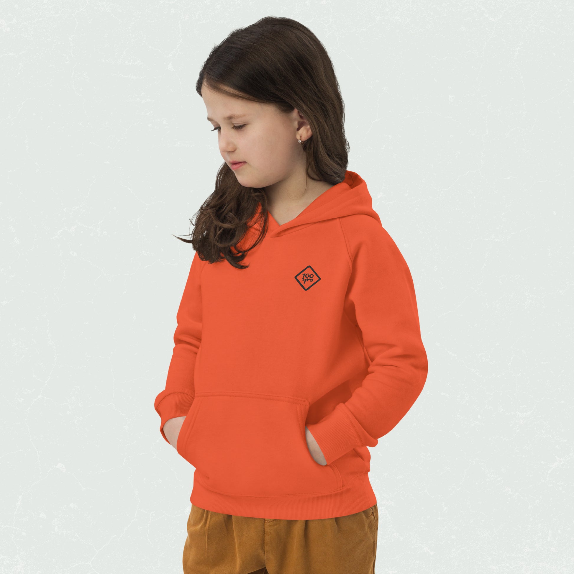 Young child wearing an orange unisex eco hoodie with 100yrs logo on the chest, hands in front pouch pocket, looking down thoughtfully.