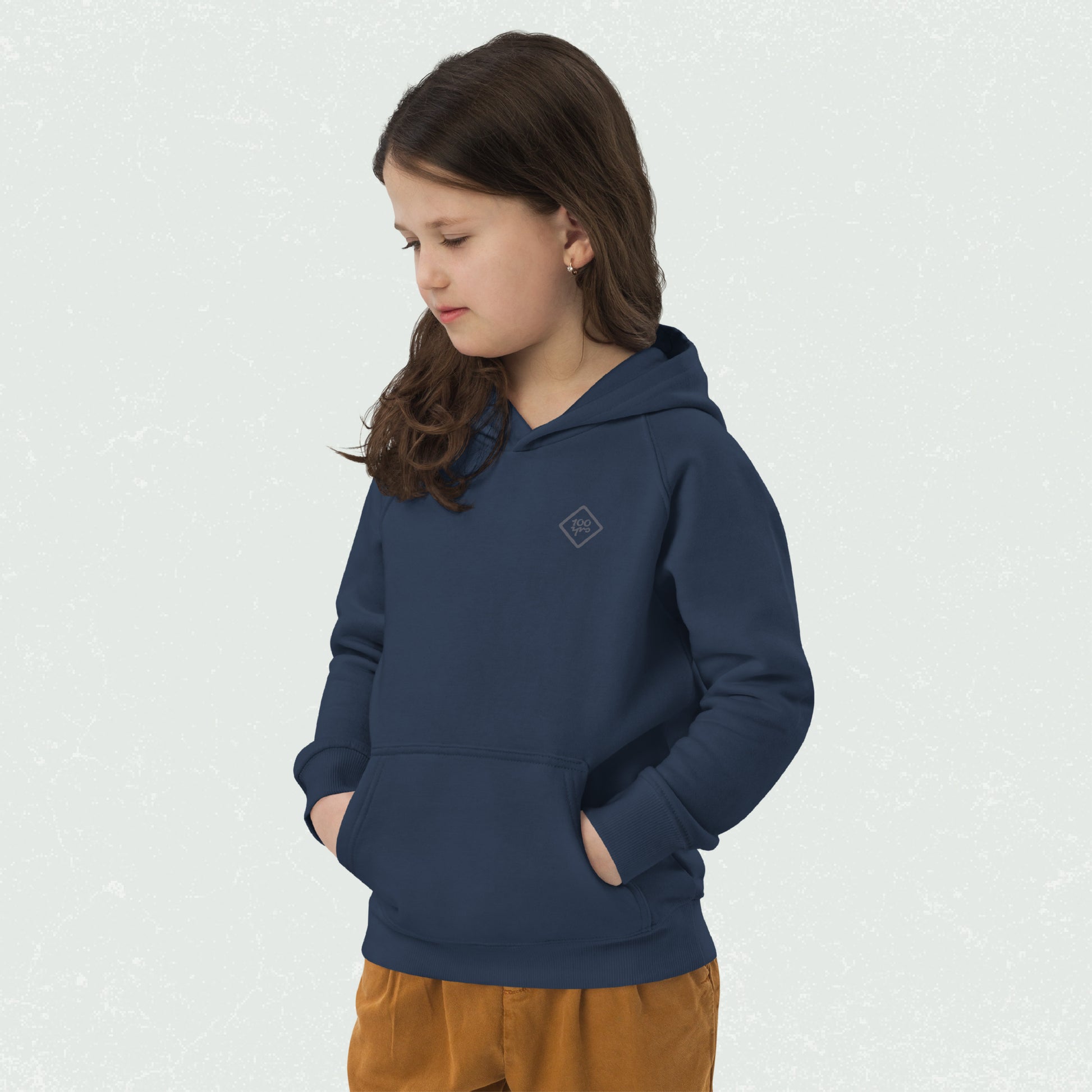 Young girl wearing a navy blue Unisex Kids Eco Hoodie with "100yrs" logo on the chest, hands in the front pouch pocket, and looking down; matched with comfy mustard yellow pants.