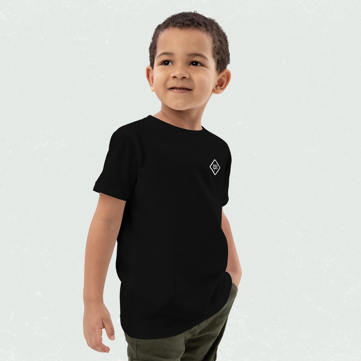 Kid wearing a black Unisex Organic Kids T-Shirt with the 100yrs logo, smiling confidently while posing with one hand in their pocket.