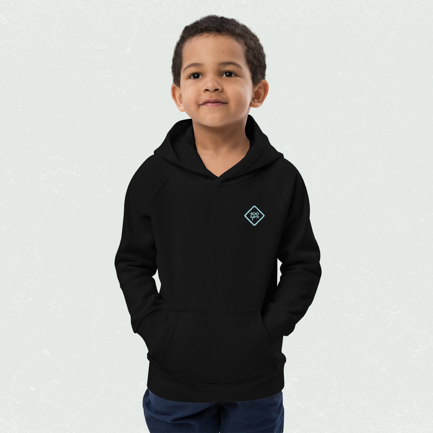 Kid wearing a black unisex eco hoodie with '100yrs' logo, hands in front pouch pocket, looking cheerful and cozy.