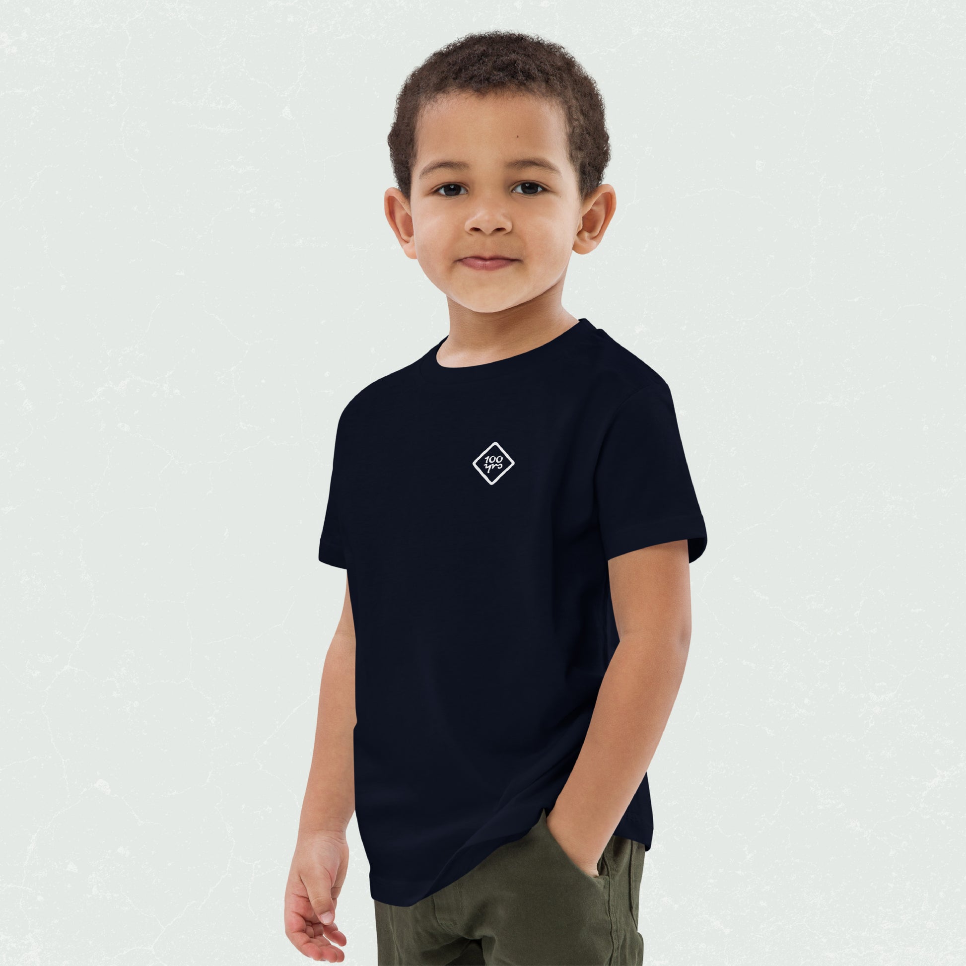 Young boy wearing a navy blue unisex organic kids t-shirt with a small white "100yrs" logo on the chest, standing with a hand in his pocket and smiling confidently.