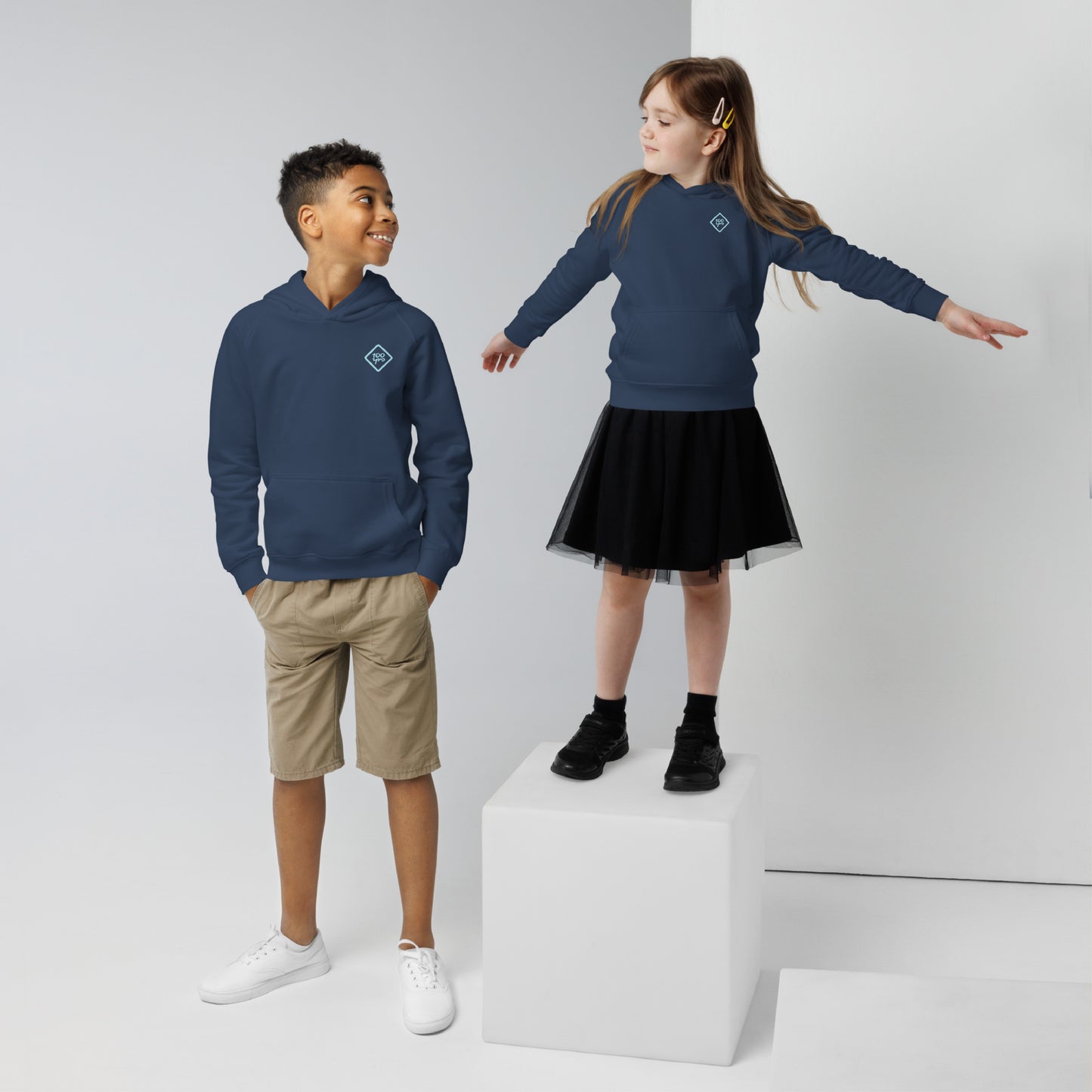 Two kids in blue unisex eco hoodies, one standing on a white block reaching out while the other smiles with hands in pockets.