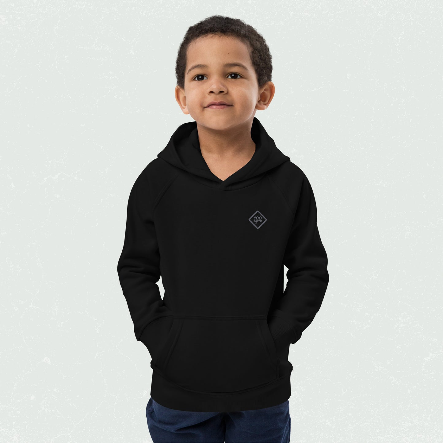 Little boy wearing a black Unisex Kids Eco Hoodie with "100yrs" logo, hands in front pocket, and a soft smile.