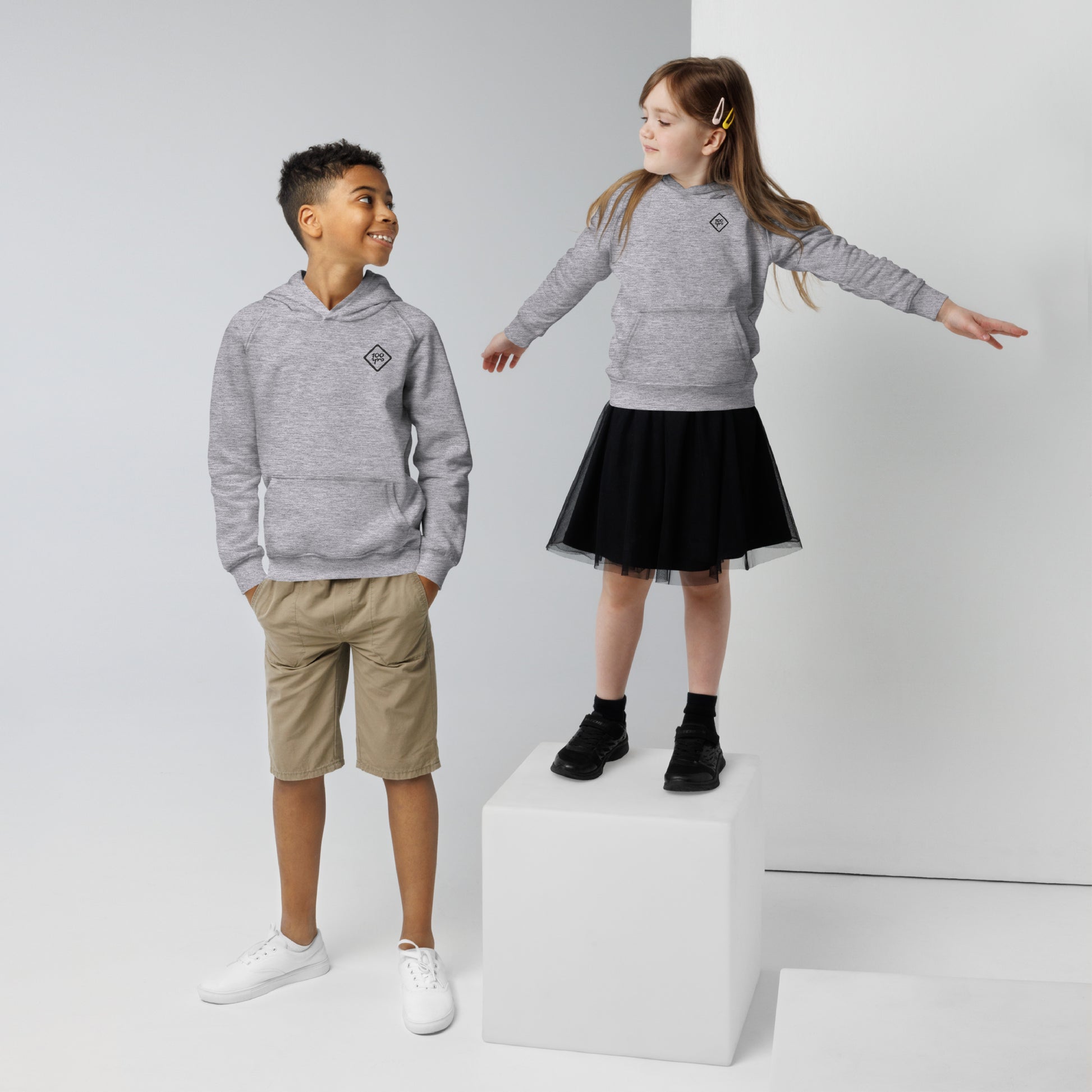 Kids wearing the Unisex Kids Eco Hoodie with the 100yrs Logo. Boy in khaki shorts and girl in a black skirt. Cozy and stylish!