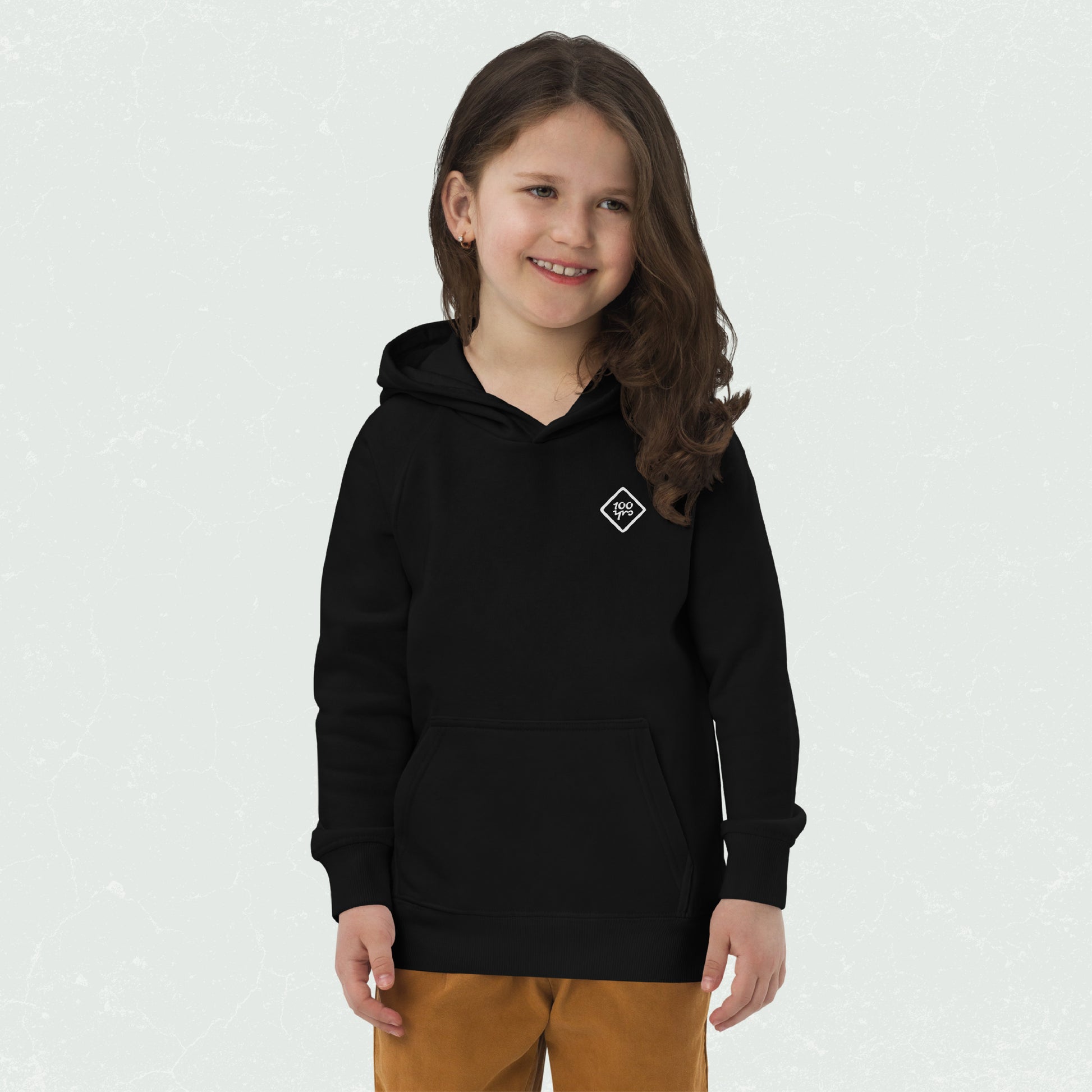 Young girl wearing a black "100yrs Logo" eco hoodie with a front pouch pocket and relaxed fit.